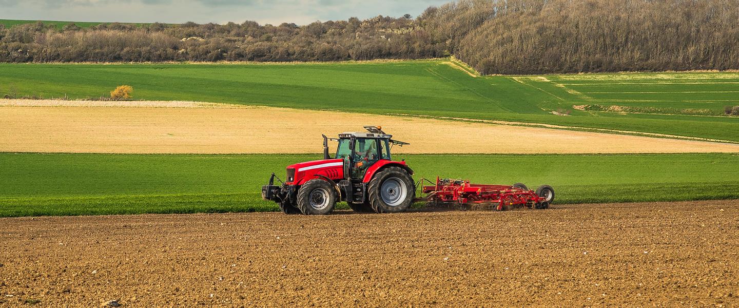 Red tractor pulling equipment through soil in field with other green farming fields in background