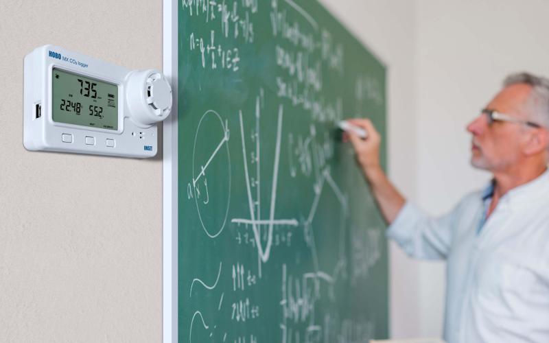 Teacher writing on a chalkboard with a HOBO MX1102A CO2 data logger mounted on the wall