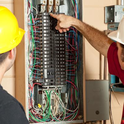 2 men in hard hats looking at an electrical panel, one of them pointing at a breaker in the panel.