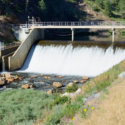 A view of a spillway with a walkway going across it, rocks and vegetation at the bottom of the falls.